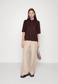 Camisa button-up de mangas curtas em tom castanho, corte descontraído, combinada com calças largas bege e sandálias de plataforma pretas, segurando uma carteira com padrão.