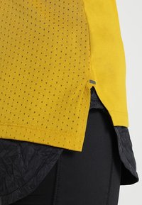 Top giallo a maniche corte con texture perforata, caratterizzato da un orlo arrotondato e un sottostrato nero per contrasto. Accentuato con una linguetta grigia.
