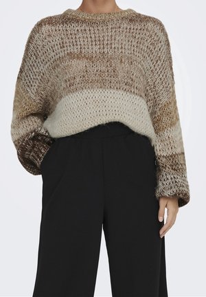 Person iført en løst strikket, flerfarvet beige og brun sweater med faldende skuldre og højttaljede sorte bukser, den ene hånd i lommen.