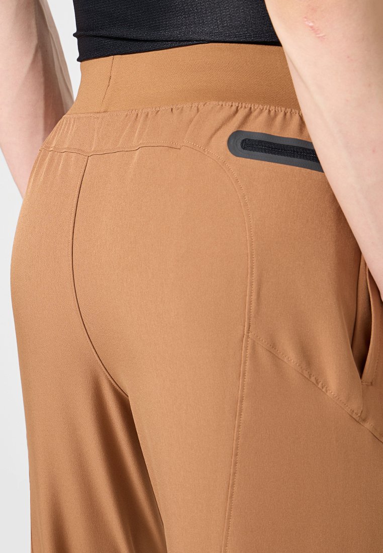Pantalones deportivos marrones con una textura suave, que presentan una cintura elástica y un bolsillo lateral con un detalle de cremallera negra.