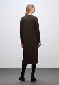 Brun langærmet kjole med lige snit, der går til midt på læggen, lavet af blødt stof. Båret med sorte ankelstøvler, minimal detaljer på ryggen.