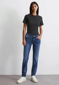 Mujer de pie contra una pared blanca lisa, vestida con una camiseta negra de manga corta, jeans ajustados azules y zapatillas blancas.