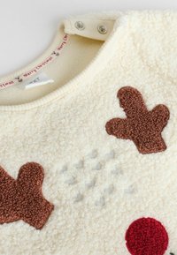 Cremfarbener Fleece-Oberteil mit braunen, plüschigen Lebkuchenformen und einem roten runden Akzent. Verfügt über einen Druckknopfverschluss am Kragen.