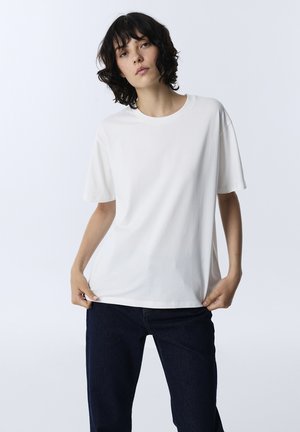 Woman with short curly dark hair wears plain white oversized t-shirt and dark jeans, holding shirt hem while standing against white background.