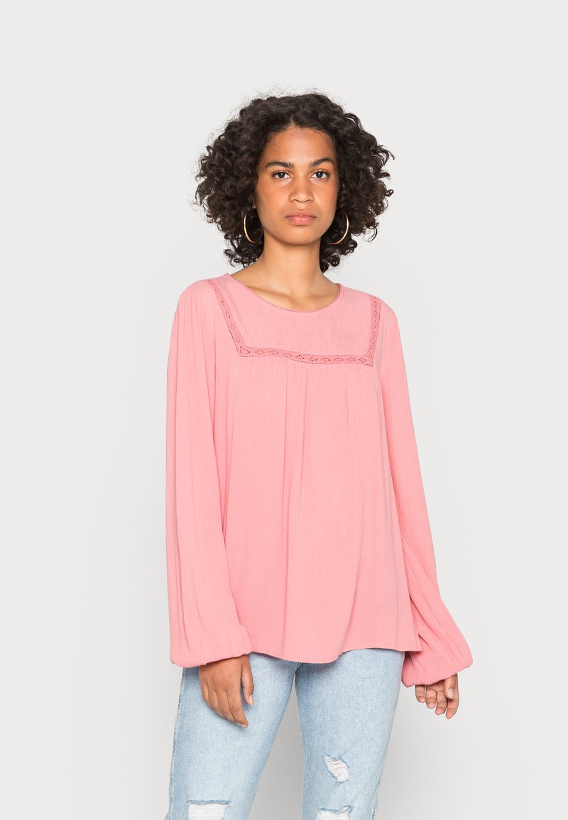 Blusa rosa de mangas longas com um tecido texturizado, apresentando um decote redondo e detalhes bordados no peito, combinada com jeans de denim claro.