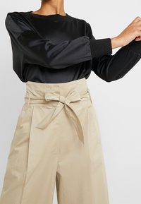 Svart långärmad blus med rynkade manschetter, tillsammans med högmidjade beige byxor knutna med en snabb vid midjan.