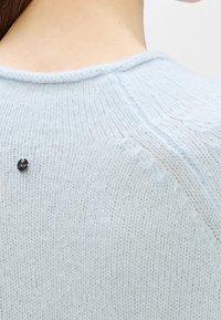 Gros plan du dos d'un pull tricoté bleu clair avec un petit bouton rond noir près de la couture d'épaule sur le cou d'une personne et les cheveux visibles.
