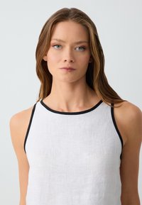 Haut sans manches en lin blanc avec des bordures noires. Design simple, col haut et texture lisse. Convient pour les temps chauds.
