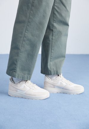Personne portant des baskets Nike beige clair et un pantalon large gris-vert, debout sur un sol bleu.