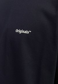 Tissu noir avec texte blanc "Originals™" imprimé, montrant une texture textile fine et de légers plis.