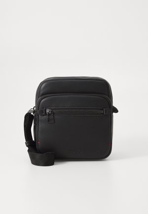 Kompakt sort crossbody-taske med frontlomme med lynlås, justerbar vævet strop og diskret mærkeprægning på glat materiale.