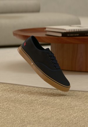 Sort snøresneaker med brun gummisål og rødt logo, svævende over et beige tekstureret tæppe nær et rundt træ- kaffebord med bøger.