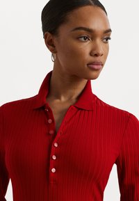 Red ribbed polo shirt with a collared neck, featuring a button placket and a fitted silhouette. Smooth fabric with a slightly textured finish.