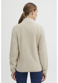 Beige fleece-genser med en myk, teksturert overflate, høy krage og lange ermer. Har en rett nederkant og en litt avslappet passform.