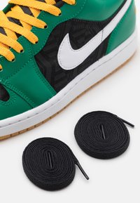 Baskets Nike vertes et noires avec des lacets jaunes à côté de deux lacets noirs enroulés sur fond blanc.