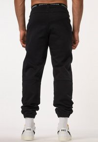 Pantalones de chándal negros con una cintura elástica que presenta una marca blanca. Corte ajustado con puños elásticos. Llevado con calcetines blancos y zapatillas.