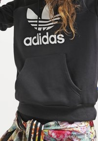 Svart hoodie med en stor vit Adidas-logotyp. Har en framficka i kengurustil och texturerat tyg. Blommönstrat midjeband syns nedanför.