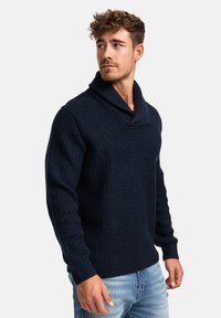 Granatowy sweter dzianinowy z kołnierzem szalowym, charakteryzujący się wzorem teksturowym. Długie rękawy i ściągacze przy mankietach oraz u dołu. Noszony z jasnoniebieskimi dżinsami.