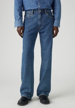 Osoba ubrana w niebieskie jeansy o prostych nogawkach, pasującą niebieską koszulę dżinsową oraz czarne buty, stojąca na jednolitym szarym tle.