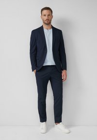 Giacca da abito blu navy con revers a punta, abbinata a una maglietta azzurra chiara e pantaloni blu navy slim-fit. Completato con sneakers bianche.