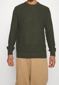 Mörkgrön stickad tröja med ett strukturerat mönster, rund halsringning och ribbade ärmslut och nederkant. Matchad med ljusbeiga byxor.