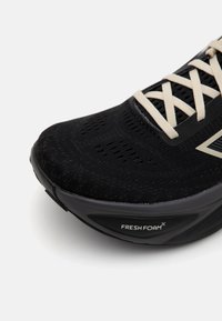 Gros plan sur une chaussure de sport noire avec des lacets beige et une semelle épaisse portant la mention Fresh Foam sur fond blanc.