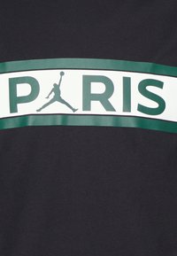 Black cotton t-shirt featuring a large white band with green text spelling "PARIS" and a black Jordan logo silhouette in mid-air.