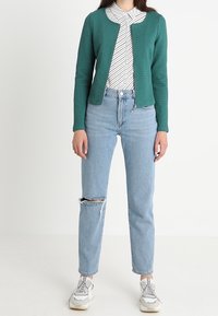 Kobieta w turkusowej, teksturowanej bluzie na zamek, w białym, paskowanym topie, jasnoniebieskich, przetartych dżinsach i białych sneakersach, stojąca na białym tle.