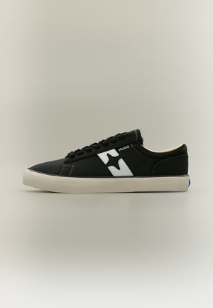 Baskets basses noires avec semelle blanche, logo Hugo blanc sur le côté et lacets noirs sur fond beige uni.