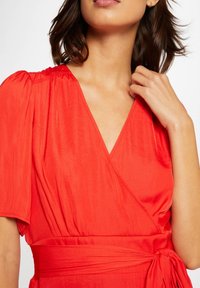 Robe portefeuille rouge avec un décolleté en V, des manches courtes bouffantes et un détail de taille nouée. Tissu fluide et lisse avec un drapé subtil.