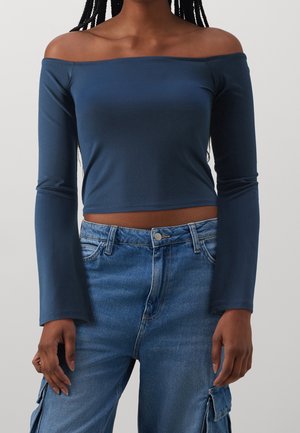 Persoon draagt een marineblauw off-shoulder crop top met lange mouwen en een hoge taille blauwe denim cargobroek, staand tegen een effen achtergrond.