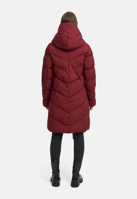 Rød polstret jakke med hette, med et chevron quiltemønster, midtlang design, og brukt med svarte leggings og støvler.