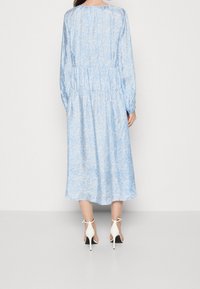 Vestido de manga larga de estampado floral en azul claro, con cintura fruncida, falda fluida y escote redondo, combinado con zapatos de tacón blanco.