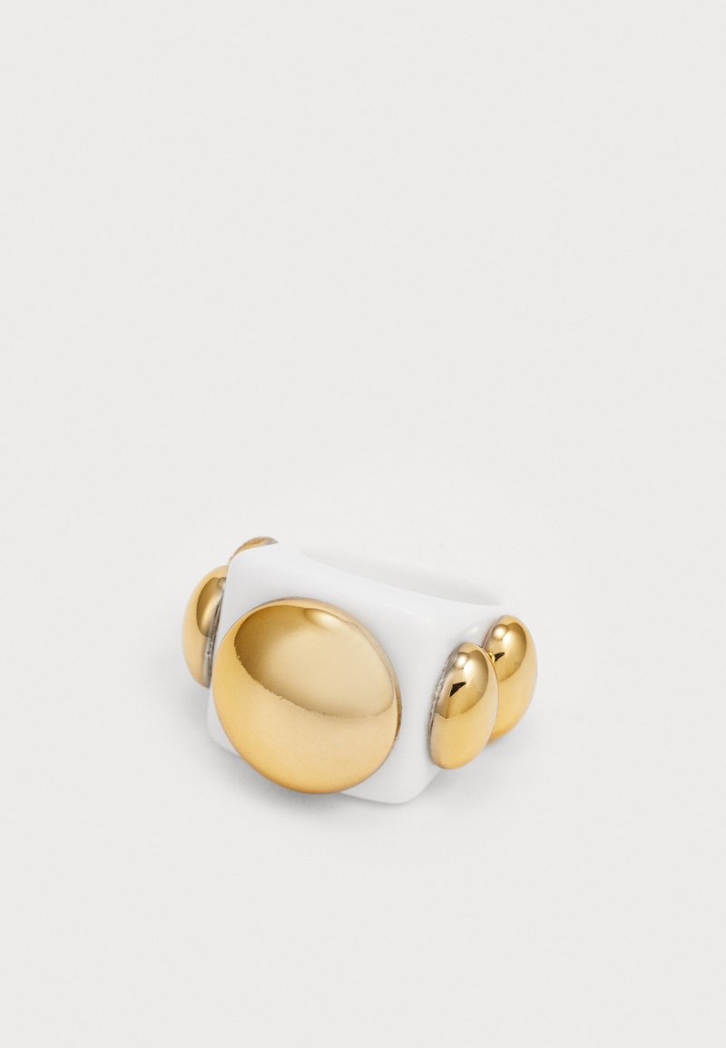 Bílý prsten s lesklým povrchem, na jedné straně s velkým zlatým kruhovým akcentem a třemi menšími zlatými kuličkami. Hladký povrch.
