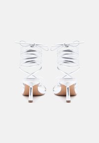 Ασημένια σανδάλια με ψηλό τακούνι και λεπτά σπειροειδή λουράκια στον αστράγαλο, δεμένα σε φιόγκους, εμφανιζόμενα από πίσω σε λευκό φόντο.