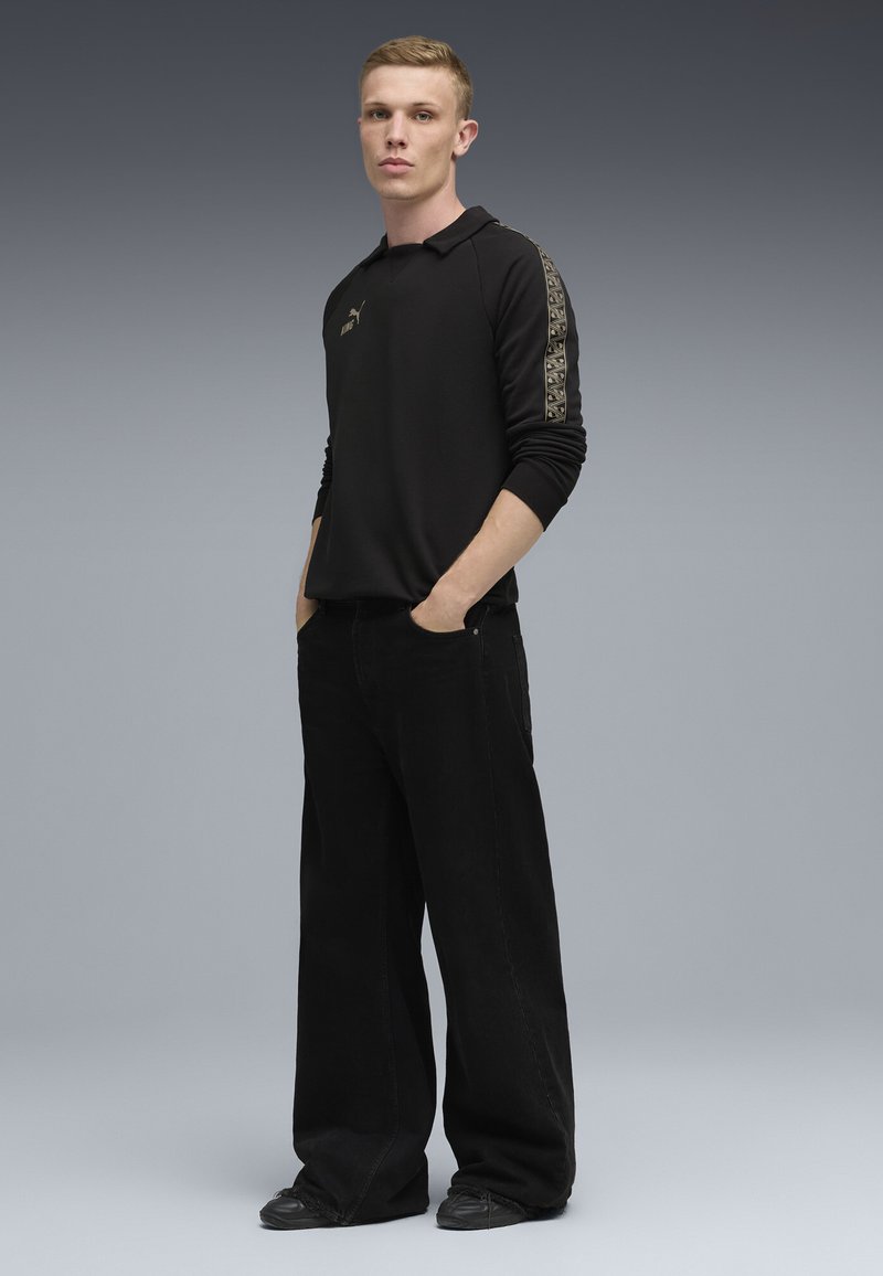 Un jeune homme se tient confiamment avec les mains dans les poches, portant un t-shirt noir à manches longues et un pantalon noir à jambe large devant un fond dégradé.