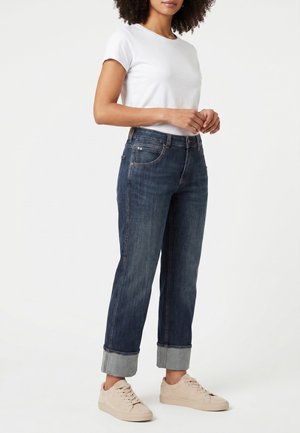 Mujer con vaqueros oscuros remangados, camiseta blanca de manga corta y zapatillas beige claras de pie frente a fondo blanco.