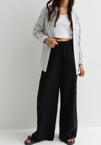 Zwarte, wijde broek van textuurstof met een elastische tailleband, gecombineerd met een witte cropped tanktop en een gestreepte blouse met lange mouwen.