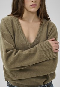 Pull oversize vert olive en tissu maille douce, avec un col en V profond, des poignets côtelés et une coupe décontractée. Design minimaliste.