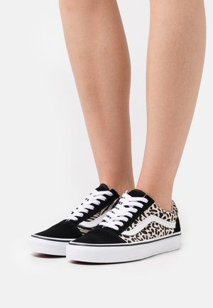 Jambes portant des baskets basses noir et blanc avec des panneaux à imprimé animal et des lacets blancs sur un fond blanc.