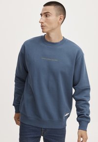 Blauwe sweatshirt gemaakt van zachte stof met een ronde hals. Het heeft gouden tekst op de borst en ribbelboorden aan de mouwen en onderkant. Gecombineerd met denim jeans.