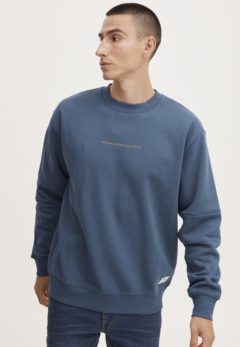 Blauwe sweatshirt gemaakt van zachte stof met een ronde hals. Het heeft gouden tekst op de borst en ribbelboorden aan de mouwen en onderkant. Gecombineerd met denim jeans.