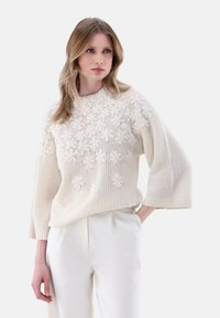 Strickpullover in Off-White mit floralen Mustern, rundem Ausschnitt und weit geschnittenen Ärmeln, kombiniert mit hellen, taillierten Hosen.
