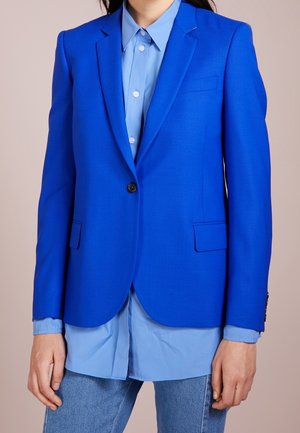 Femme portant un blazer bleu vif sur une chemise à boutons bleu clair et un jean bleu, debout devant un fond neutre.