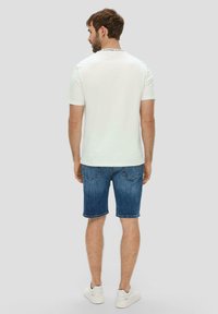 Biała koszulka z krótkim rękawem z czarną metką przy kołnierzu, w połączeniu z niebieskimi szortami dżinsowymi i białymi trampkami, pokazana z tyłu.