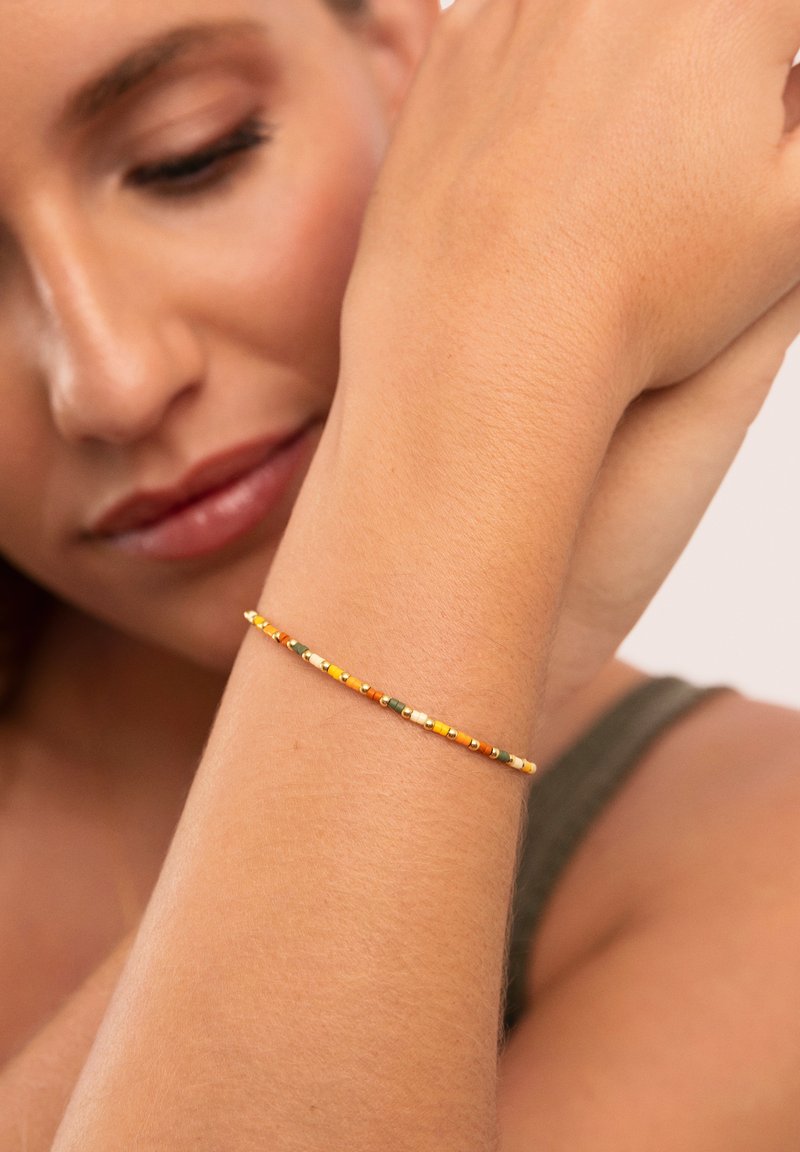 Pulsera delgada de cuentas que presenta una mezcla de cuentas en naranja, amarillo, verde y blanco. Llevada en una muñeca sobre un fondo neutro.