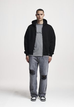 Nuori mies seisoo kasvot eteenpäin, yllään musta vetoketjullinen huppari, harmaa graafinen t-paita, revityt harmaat farkut ja mustavalkoiset tennarit yksivärisellä taustalla.