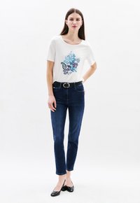 Witte kortetermijn T-shirt met blauwe bloemenborduring, gecombineerd met donkerblauwe slim-fit jeans en zwarte puntige flats.