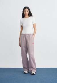 Γυναίκα με μακριά σκούρα μαλλιά στέκεται σε μπλε πάτωμα, φοράει εφαρμοστό λευκό κοντομάνικο πουκάμισο, φαρδιά λιλα παντελόνια φόρμας και λευκά αθλητικά παπούτσια.