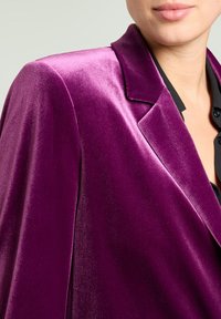 Blazer de terciopelo púrpura con cuello estructurado, que presenta una textura suave y líneas nítidas, superpuesto sobre una camisa negra.